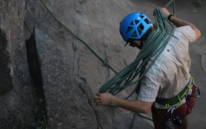 A person wearing safety gear uses their shoulders to fold a rock climbing rope while standing next to a rock wall